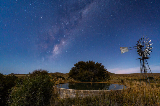 Stunning night photograph of a traditional wind pump or windmill and reservoir under the vibrant Milky Way in the arid Great Karoo, Western Cape, South Africa. Serene rural landscape with starry skies