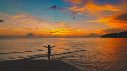 Silhouette of a young woman standing on a beach with open arms, savoring the vibrant hues of a stunning sunset over the tranquil sea in Thailand, Asia