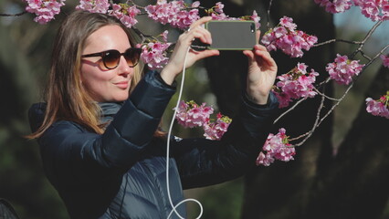 Young woman is taking pictures of beautiful cherry blossoms with her smartphone in a park in tokyo. She is enjoying the beautiful spring day
