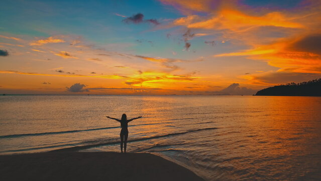 Silhouette of a young woman standing on a beach with open arms, savoring the vibrant hues of a stunning sunset over the tranquil sea in Thailand, Asia