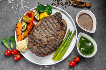 Grilled steak with veggies. A large grilled steak served with assorted fresh vegetables and sauces on a white plate, creating a vibrant meal.
