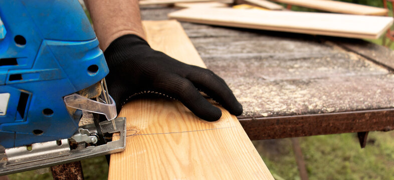 Close up of male hands of carpenter working with electric jigsaw in protective gloves cutting wood with saw, woodworking hobby concept