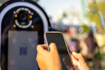 Close-up of hands holding a smartphone in front of a modern photo booth with camera lens and QR code scanner during an outdoor public event in daylight.