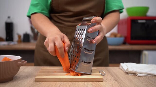 Woman in apron grating carrot in glass bowl at table in kitchen. Vegan food and healthy diet. Cooking vegetables salad