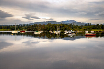 Obraz premium yachts and boats reflected in the still, calm waters of a huge lake with distant trees and mountains, Lake Windermere, Cumbria, The Lake District, England 
