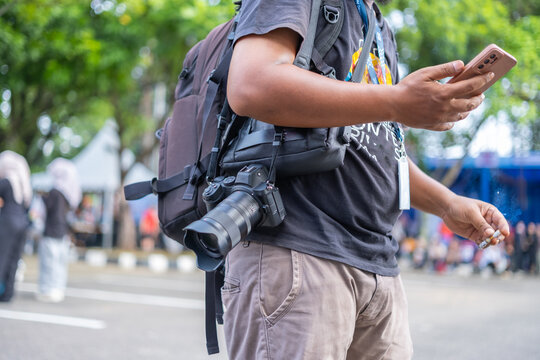 A man with a backpack and camera smokes while checking his smartphone during an outdoor street event in the city.