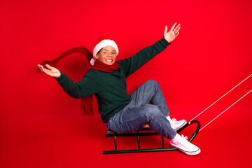 Obraz premium Festive boy in Santa hat sitting on sled, celebrating Christmas on vibrant red background with joy and magic