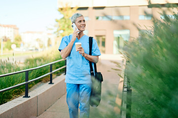 Nurse in scrubs enjoying coffee and using a mobile phone outside a modern medical facility during a sunny day