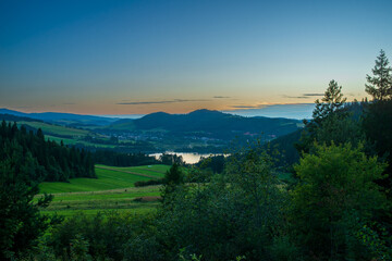 Green meadows and dark forests surround the shimmering Czorsztyn Lake as the last light of day fades. The panoramic view captures the quiet beauty of southern Poland, ideal for eco-tourism and rural t