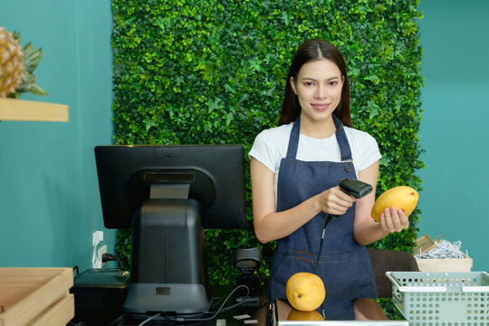 Caucasian female fruit seller smiling holding barcode scanner and ripe mango behind checkout preparing front counter during early business setup inside modern organized grocery store before opening