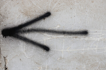 A close-up view of a black spray-painted arrow on a textured concrete wall, indicating direction with two diverging lines. The background features light scratches and marks, enhancing the urban