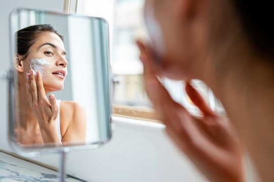 Woman Smiling in Mirror While Applying Skincare Cream - Powered by Adobe