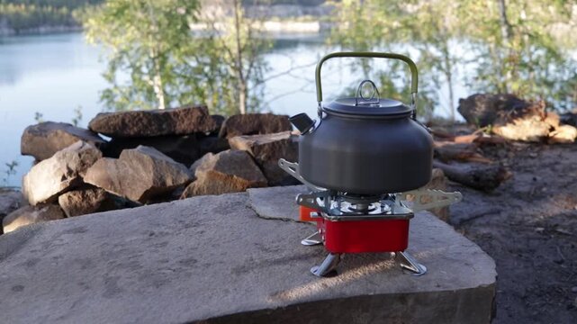 Kettle on a portable gas stove in a scenic forest landscape by the lake. Cozy camping atmosphere, outdoor cooking, and relaxation in nature.