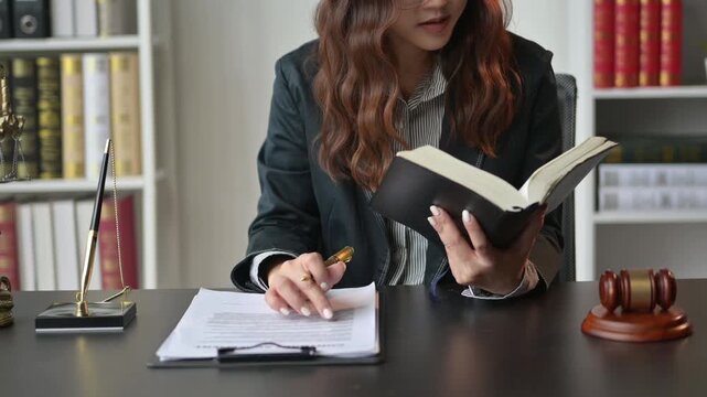 woman in a suit is reading a book while sitting at a desk. She is holding a pen and a clipboard. The scene suggests a professional setting, possibly in a law office or a courtroom