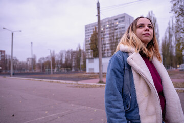 Fototapeta premium Young woman in casual attire stands in an urban park during early evening, surrounded by tall buildings and autumn trees
