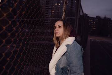 A young woman stands near a chain-link fence in an urban area during the late afternoon of an autumn day