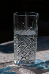 An elegant cut-crystal glass filled with water sits on a sunlit table. The intricate pattern on the glass refracts light, creating a sparkling effect that suggests refreshment and classic refinement. 