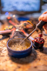A close-up view of a soldering iron being used to work on electronic FPV components, showcasing a tin of solder and a motor on a workbench. neutral background, clear negative space, clean composition