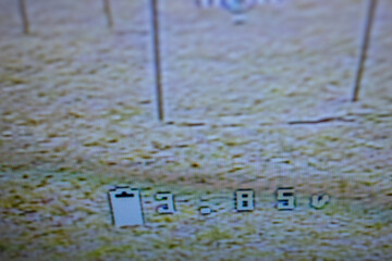 A close-up view of a digital display showing battery status of FPV Drone and other information on a screen, with a blurred background of a grassy field. neutral background