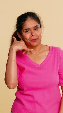 Indian woman playfully forms call me gesture by placing hand near ear, smiling brightly. Hindu girl on beige background happily suggesting phone call, communication, casual chat, or friendly contact.