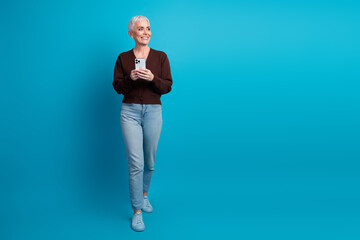 Smiling mature woman holding smartphone against blue background, wearing casual cardigan and jeans, trendy and stylish