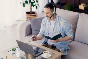 Handsome man with headset using a laptop and book for a remote professional session at home in a cozy setting