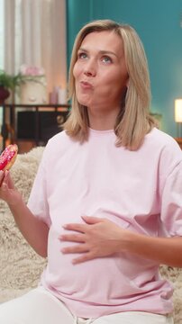 Pregnant middle-aged woman at home eats donut with appetite enjoying sugary treat craving flavor during pregnancy. Caucasian girl on sofa rolls eyes in joy showing playful mood loving fun sweet snack.