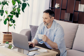 Mature professional working on laptop while relaxing at home with coffee in a contemporary and cozy living space.