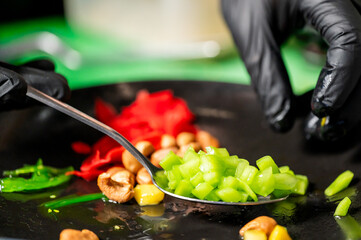 Chef plating assorted fresh ingredients on a black dish: mango, seaweed, pickled ginger, cashews, and diced vegetables. Close-up in kitchen setting, hygiene gloves visible