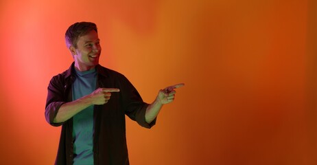 Smiling man pointing at something in neon lights against orange background. Space for text