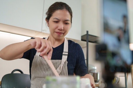 Asian woman mixes ingredients while streaming cooking vlog at home kitchen, smiling as she shares healthy homemade recipes with her audience.