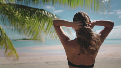 Woman standing gracefully on a stunning beach, adjusting her hair, enjoying the ocean view. Tropical setting with warm sun, clear sky, palm trees swaying