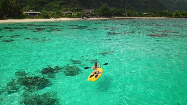 Paddling a yellow kayak in turquoise waters towards a tropical resort, a woman enjoys nature's beauty, blending adventure and relaxation in a serene lagoon