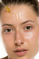 Studio shot of young woman applying serum to forehead with dropper on white background.