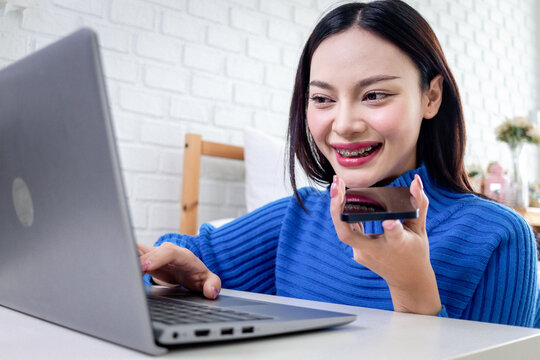 Beautiful Asian woman talking on smartphone during using laptop computer to surf on the internet. Happy young girl working online on desk in bedroom. People connect with AI technology. - Powered by Adobe