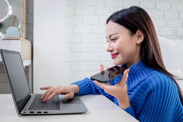 Beautiful Asian woman talking on smartphone during using laptop computer to surf on the internet. Happy young girl working online on desk in bedroom. People connect with AI technology.