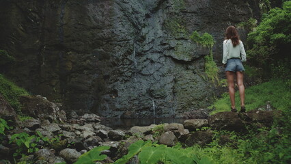 Naklejka premium Hiking woman in tropical rainforest mesmerized by waterfall on mossy rock, surrounded by lush greenery, creating peaceful moment in untouched paradise
