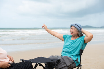 Fototapeta premium Asian senior female in casual enjoying sitting folding chair relaxing on beach seaside. Happy retired women vacation holiday lifestyle outdoor on summer.
