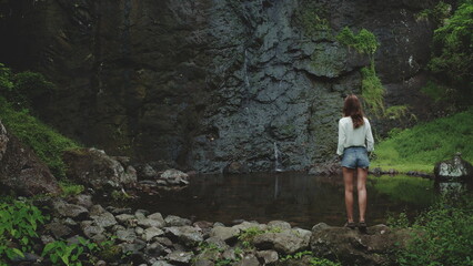 A lone woman stands on a rocky shore, admiring a cascading waterfall in the lush tropical landscape of French Polynesia. The water flows into a tranquil pool surrounded by verdant vegetation. © Anastasia Pro