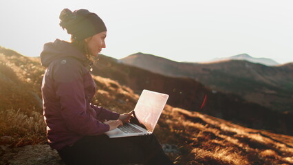 Autumn sunset mountain woman remote working on laptop. Tourist girl typing on notebook sitting on orange grass meadow in soft sun light. Hiking, trekking, outdoor lifestyle travel on holiday vacation