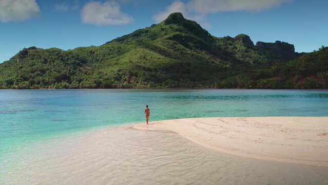 Woman wearing a bikini is enjoying a peaceful walk on the beach of a tropical island with lush green mountains in the background