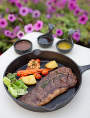 Beef Steak, A large steak on a cast iron skillet with salad greens, carrots, cherry tomatoes, and roasted potatoes or pineapple, served with steak sauce.