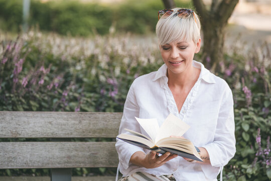 Happy adult woman reading book while sitting, outdoors - outside. Cheerful pretty adult woman with a book