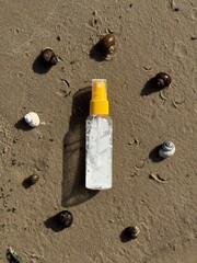 A white plastic spray bottle with a yellow cap placed on wet sand, surrounded by seashells. Minimalist beach-themed composition, suitable for concepts of summer, skincare, sunscreen, vacation
