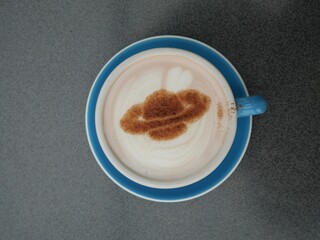 Cup of coffee with Saturn and heart foam art latte  dusted with cinnamon on blue saucer, placed on grey textured surface in South Island, New Zealand.