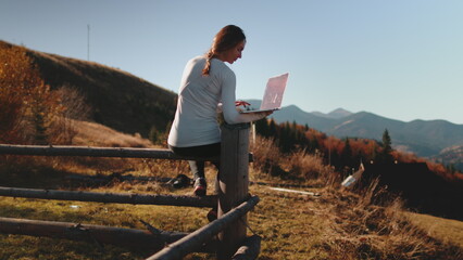 Woman works on her laptop while sitting on a mountain fence, showcasing remote work and the digital nomad lifestyle
