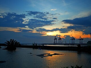 Silhouette of four port cranes at sunset with dramatic orange-red sky and scattered clouds. Industrial harbor scene during golden hour.