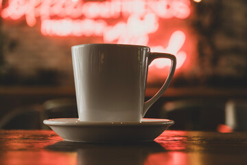 Modern cafe concept with coffee cup and neon glow. White coffee cup with neon sign background. Hot coffee in front of blurred neon lights. Trendy coffee cup with vibrant neon background.