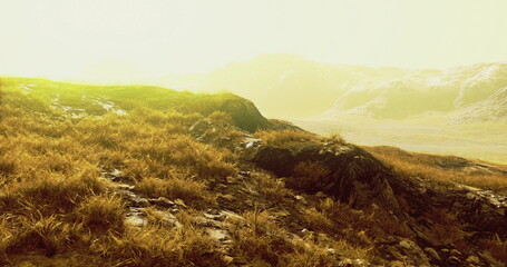 Warm sunlight filters through a hazy atmosphere, illuminating grassy hills and rocky formations. The scene captures the serene beauty of nature at dawn, inviting exploration and tranquility.