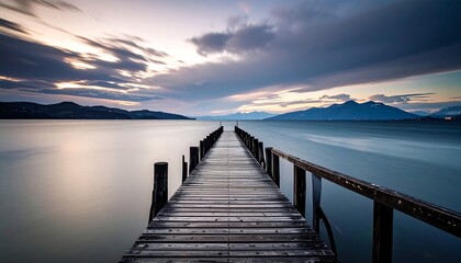 Fototapeta premium A tranquil wooden pier extends into a serene lake at dusk, reflecting the soft light of a cloudy sky.